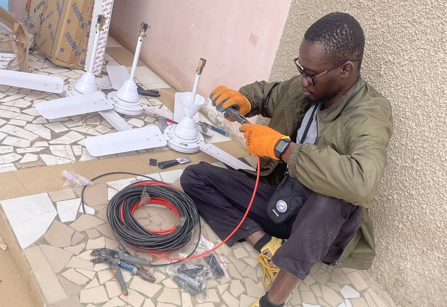 Un technicien assis au sol prépare des câbles solaires, à côté de ventilateurs de plafond à installer