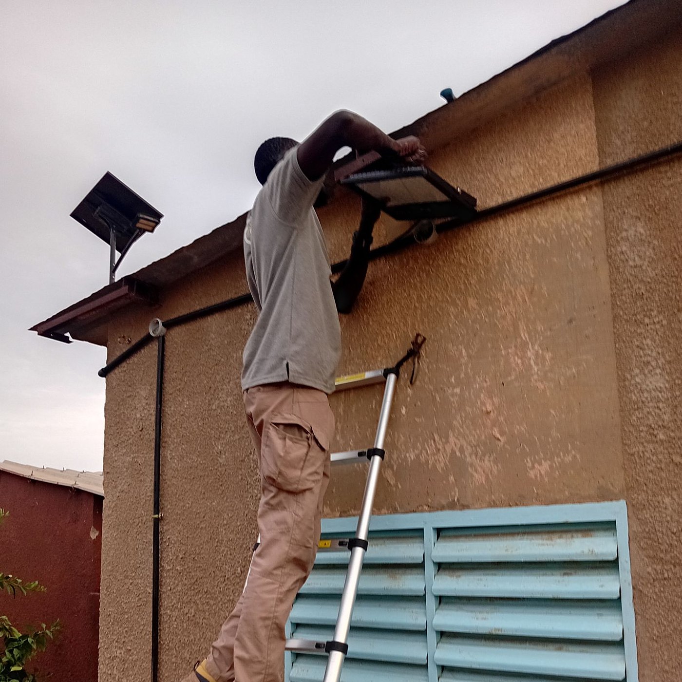 Un technicien, sur une échelle, fixe un projecteur solaire sous le toit d’un poste de santé